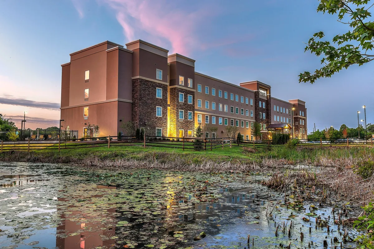 This image displays the hotel building set beside a reflective pond, surrounded by lush greenery and under a sky brushed with sunset hues. A walking path around the pond invites guests to step outside and explore, evoking a sense of tranquility while being conveniently close to the hotel.