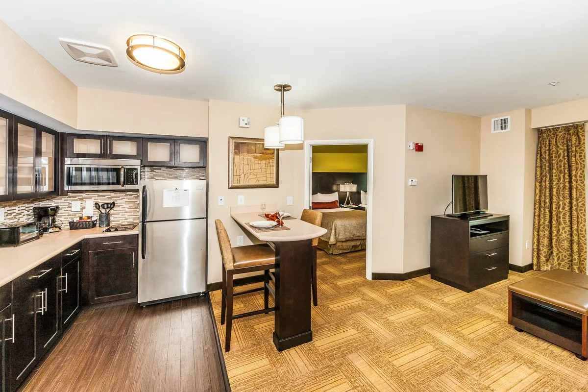 The image shows an open-plan living area with a kitchen, breakfast bar, visible living space, and the entrance to a bedroom. The living area appears comfortable and modern, with a pull-out sofa and space for guests to relax. The layout showcases the suite's functionality and how the living and sleeping areas connect, ideal for families or extended stays.