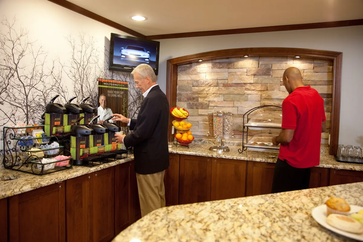 This interior image focuses on a well-appointed breakfast area, showcasing a coffee station, a colorful fruit display, and a tempting array of pastries. The space is lively yet inviting, with guests interacting and enjoying their morning meal together.