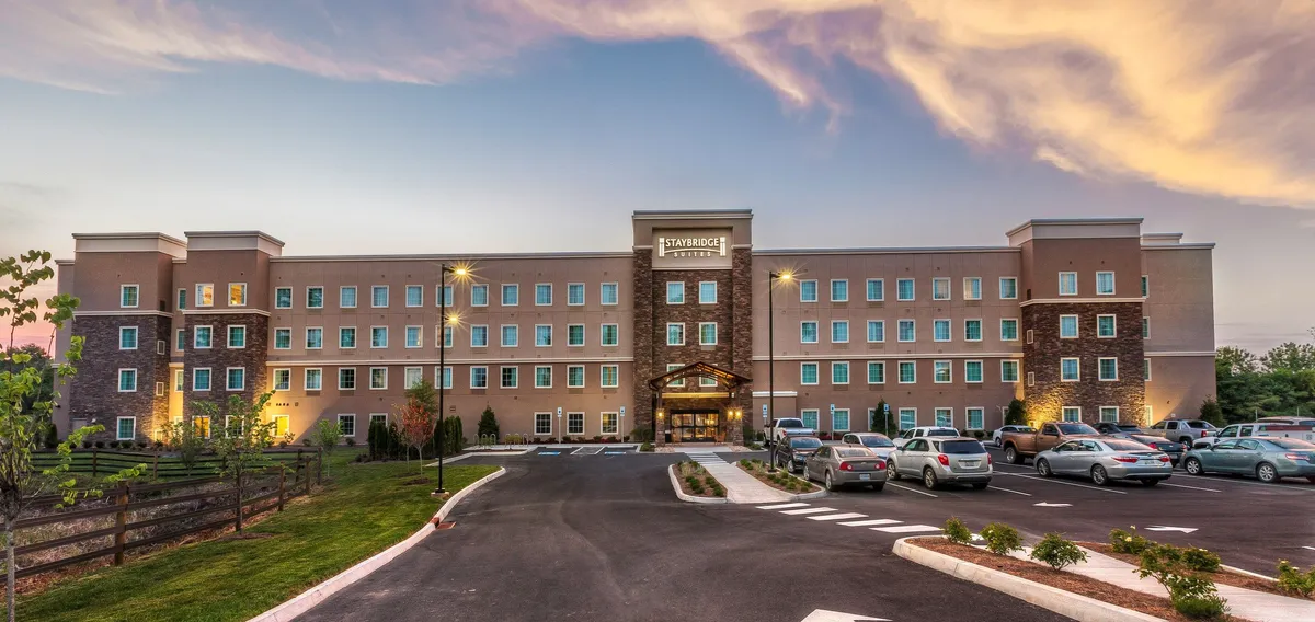 The exterior of the hotel is pictured with a well-lit entrance, landscaped greenery, and a visible, spacious parking lot. The mood is tranquil and inviting, with sunset hues adding warmth to the scene.