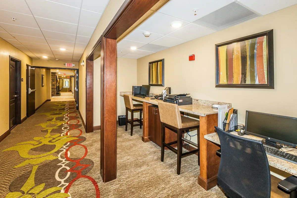 A row of computer stations set against colorful wall art and a vibrant carpet, offering a productive yet inviting workspace. This setup is ideal for guests needing to stay connected and productive, whether for remote work or personal projects, with plenty of seating and natural light.